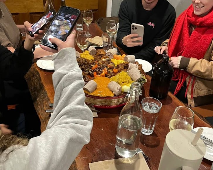 Four people sit at a restaurant table, and are taking pictures on their phone of the giant dish of Ethiopian food in front of them, It is a big share platter, and they are smiling.