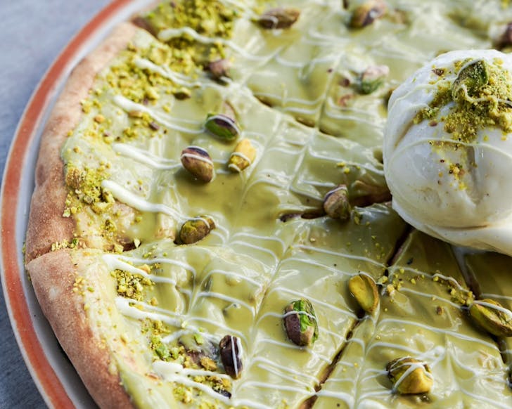 An extreme close up of a pizza with creamy green pistachio spread on top. There are decorative pistachio nuts and white chocolate drizzle all over the top, and the edge of a scoop of vanilla gelato can be seen on the middle of the pizza.