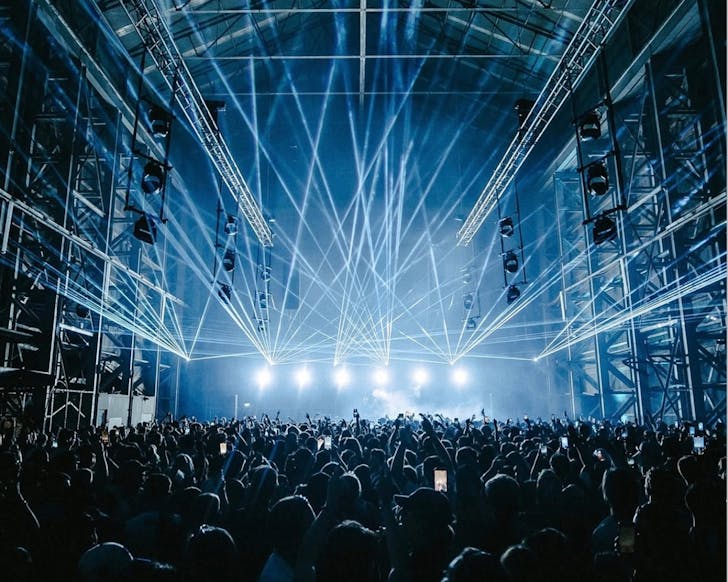 A wide shot of an indoor electronic music festival, with a large crowd of people dancing while strobe lights scatter around the room from the stage.