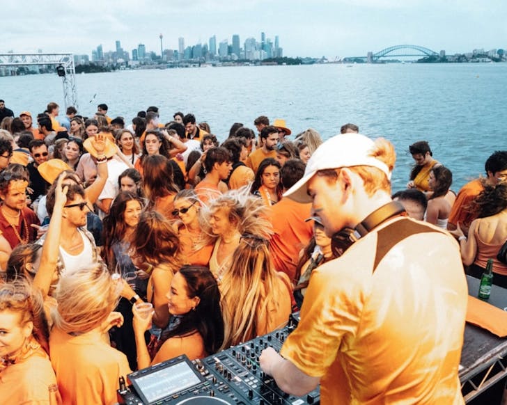 A wide shot from a boat showing a DJ facing a crowd of people dancing. Behind them and the boat is the Sydney Harbour including views of the Sydney Harbour Bridge during the late daytime.