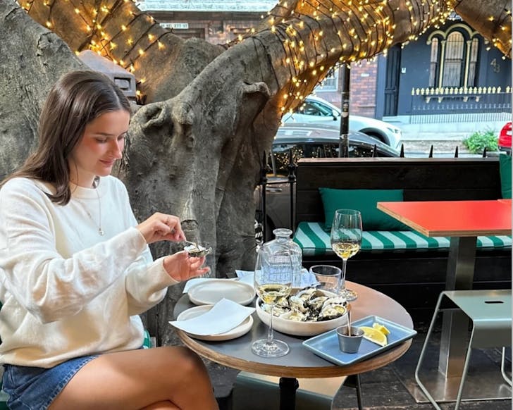 A mid shot of a lady sitting outdoors at Arms Length Bar eating oysters while the fairy lights on the tree behind her shine.