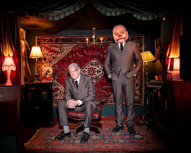 Two people in a lavish room, one wears a mask on his face.