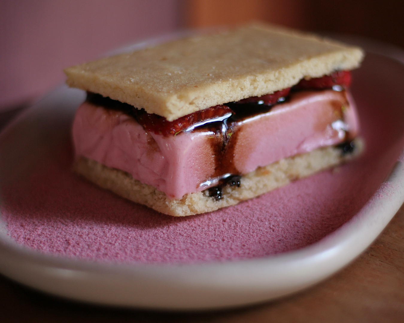 A slab of pink ice cream sandwiched between two almond cookies sitting on a dusting of pink.