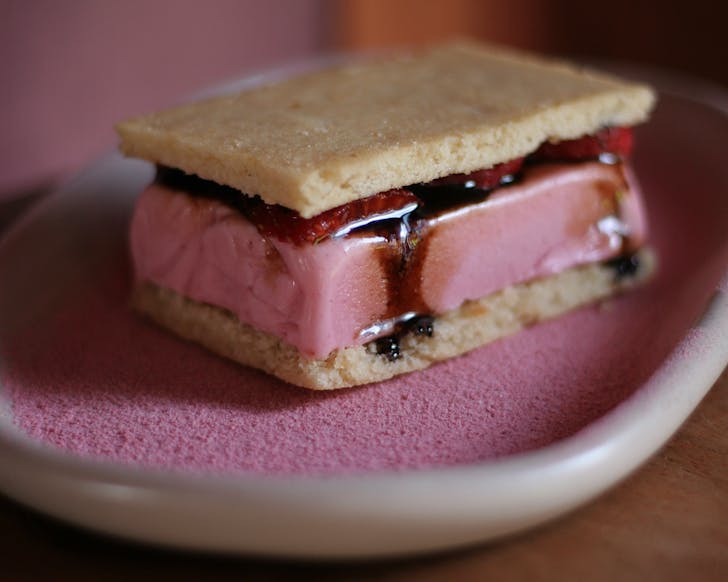 A slab of pink ice cream sandwiched between two almond cookies sitting on a dusting of pink.