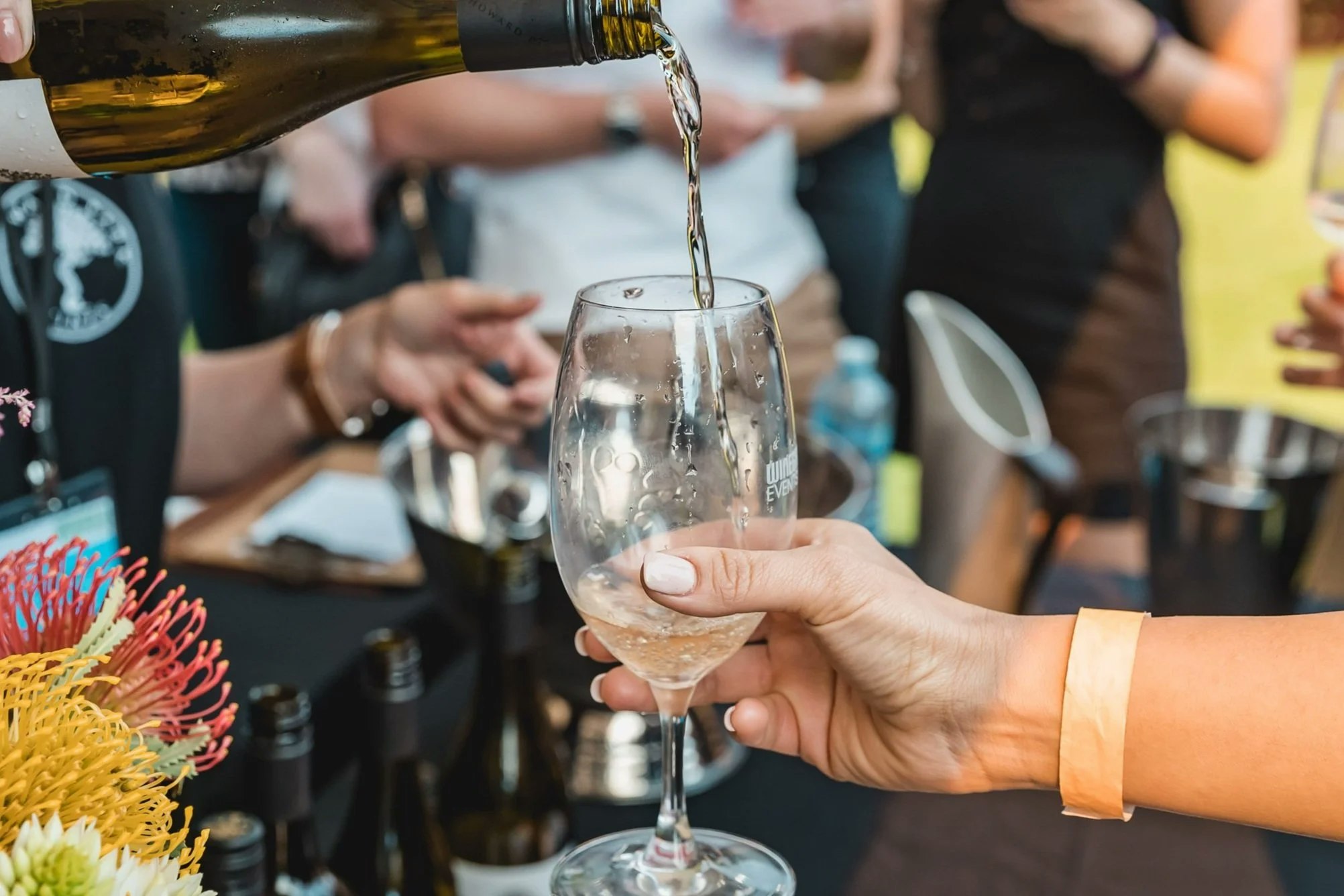 A close-up of white wine being poured into a clear tasting glass held by a person wearing a paper wristband. The scene takes place at a wine event, with bottles, flowers, and blurred people in the background, creating a lively social atmosphere.