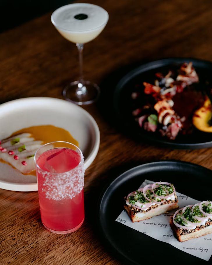 Bright pink cocktail, kingfish ceviche on black plate, sliced steak with jus on black plate, cream coloured cocktail in a martini glass on a dark wooden table