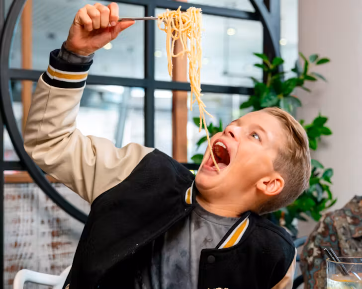 Young blonde haired Caucasian boy dangling fork filled with white butter sauce spaghetti into his mouth from high above.