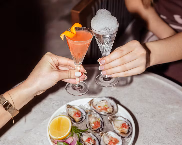 Mini orange martini with orange rind on top cheersing with mini plain martini with white foam on top, above a plate of oysters Kilpatrick.