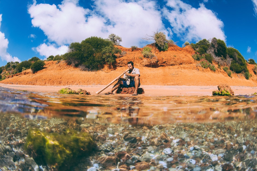 An Indigenous man sits on a golden beach while playing the didgeridoo in the sunshine.