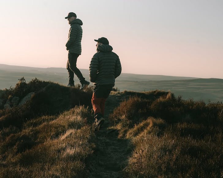 Two men wear dark green puffer jackets and wander along a ridgeline.