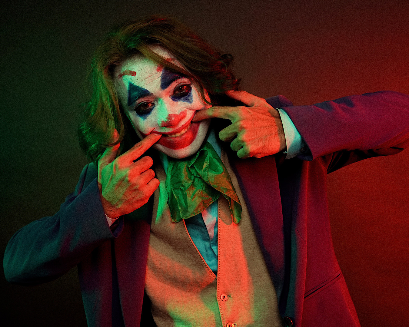 A person dressed in a Joker Halloween costume puts their fingers in each corner of their mouth pulling it into a smile. 