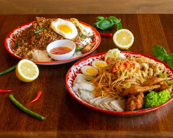 Two dishes with som tum green papaya salad and pork pad krapao with fried eggs on a wooden table decorated with lemon, chillies and fresh basil