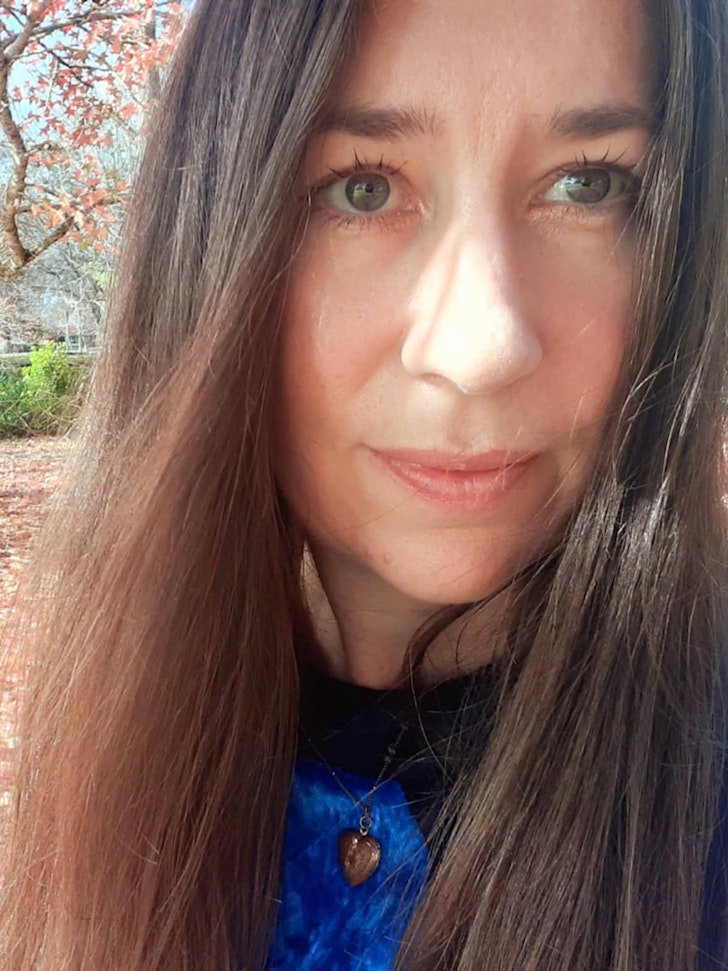 Kathryn has long dark hair and is standing in an autumnal park. 