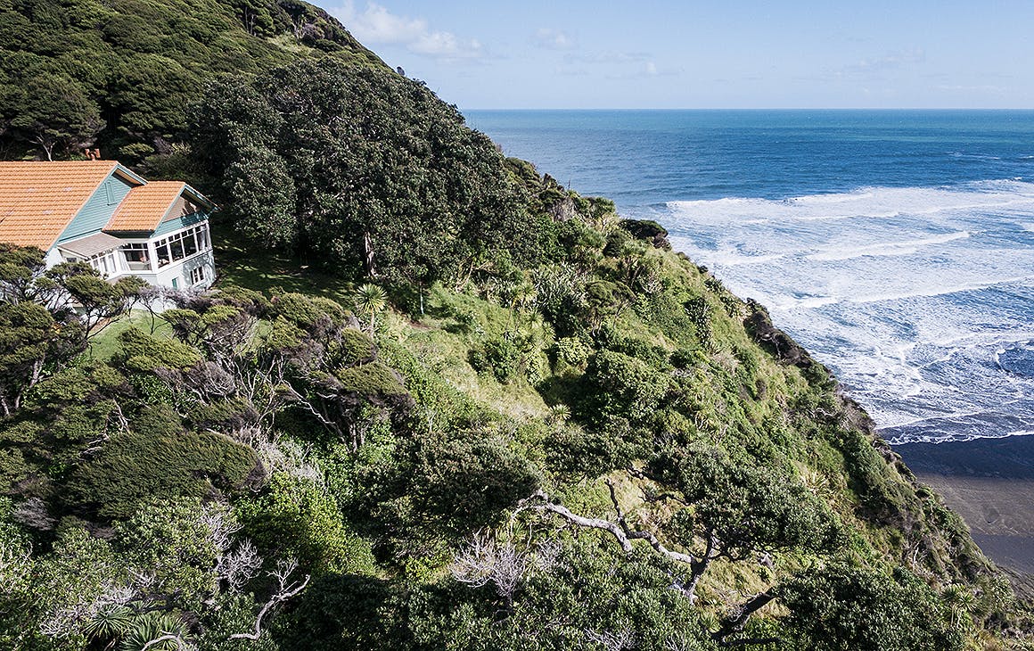 An orange-roofed 1930s heritage house sits on the side of a steep tree-covered hill, overlooking a bright blue seascape. 