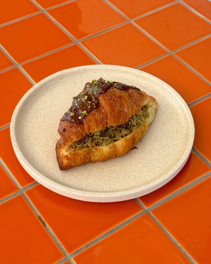 Pistachio and kataifi stuffed croissant coated in chocolate and a sprinkle of pistachios, sitting on a beige clay plate on a bright orange tiled table