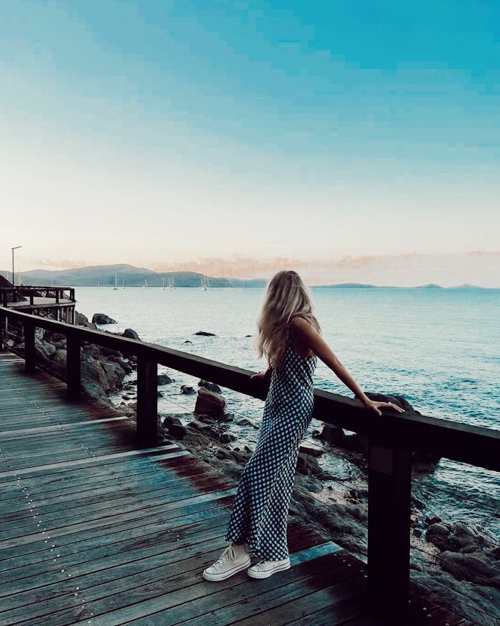 A woman standing on a boardwalk wears a gingham dress and white converse while looking out to sea. 