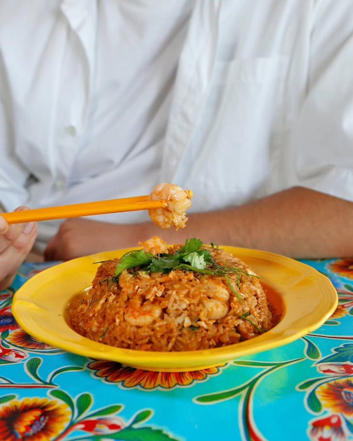 A bowl of prawn fried rice with a person in the background holding a piece of prawn with chopsticks