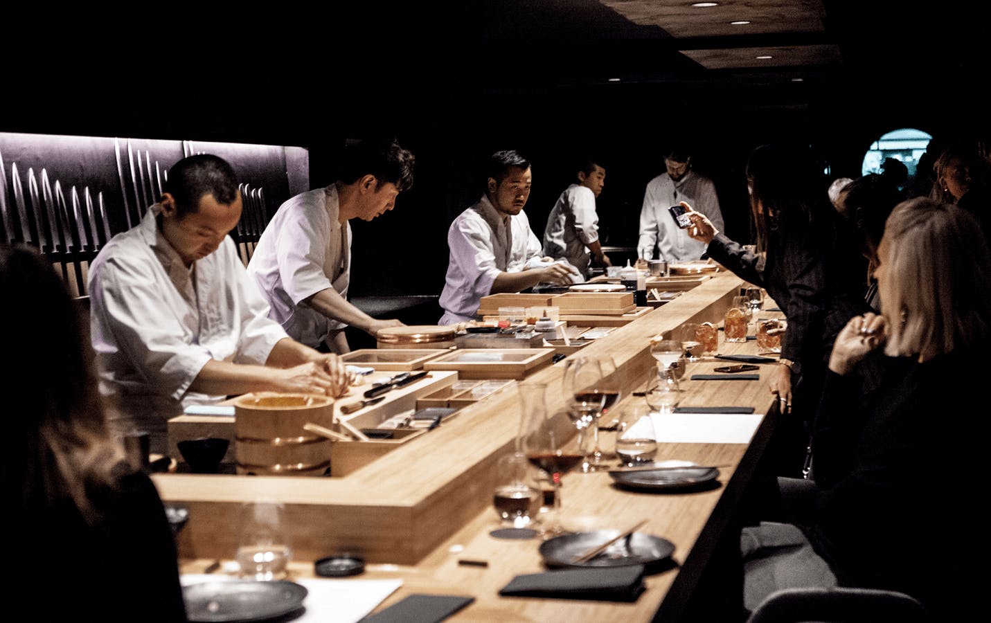 A group of chefs crafting sushi at a wooden bar with diners sitting watching. 