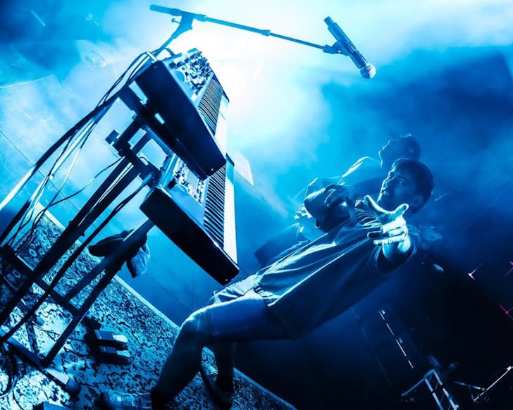 The keyboardist from L.A.B. points at the camera while performing on a blue-lighted, smoky stage.