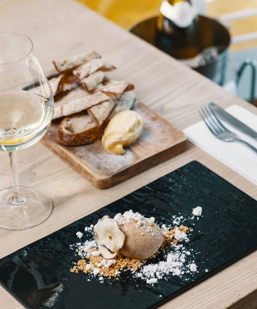 Wooden table with white wine, serving platter of sliced sourdough and beautiful presentation of gelato sitting on black plate with assortment of crushed biscuit