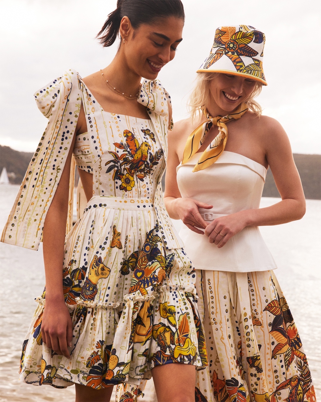 Two women wear storybook-reminiscent dresses and are smiling at the ground. 