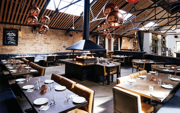 The interior at La Zeppa shows a central fireplace surrounded by chairs and tables in lush setting.