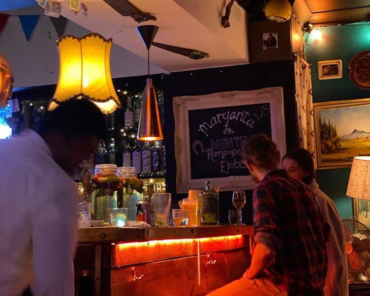 A side view of an indoor bar with low-lit lamps and decorations like a chalk board with drink specials and fake money hanging from the ceiling tastefully. A man and woman sit talking at the bar, the area is full of people and the bar table has many glasses of drinks.