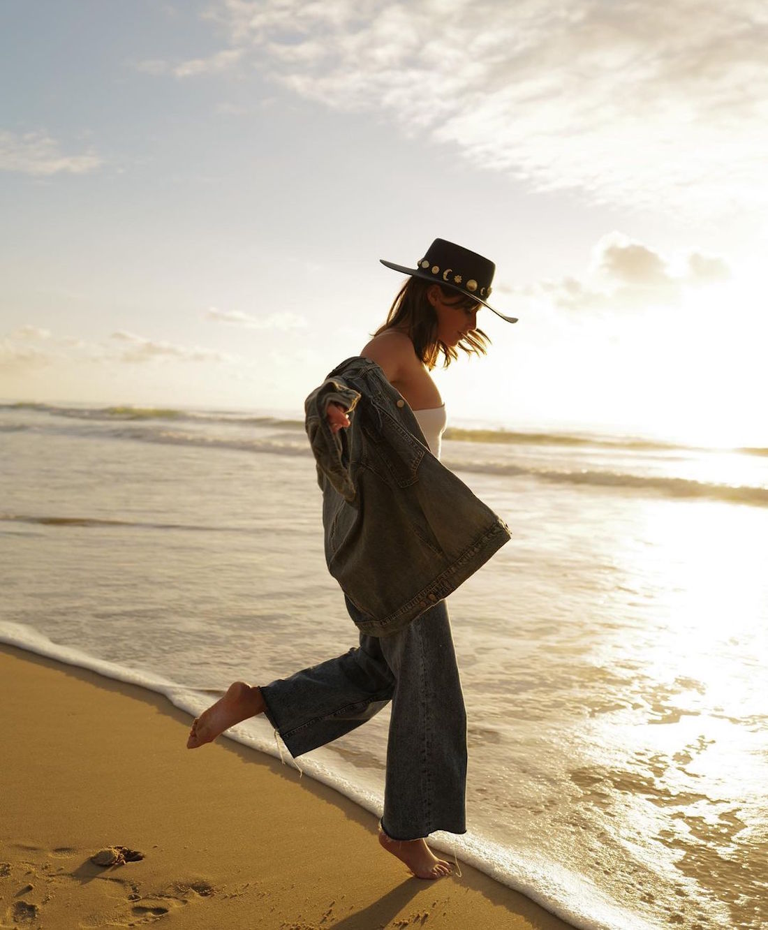 A woman walks on the beach wearing her Lack Of Colours hat.