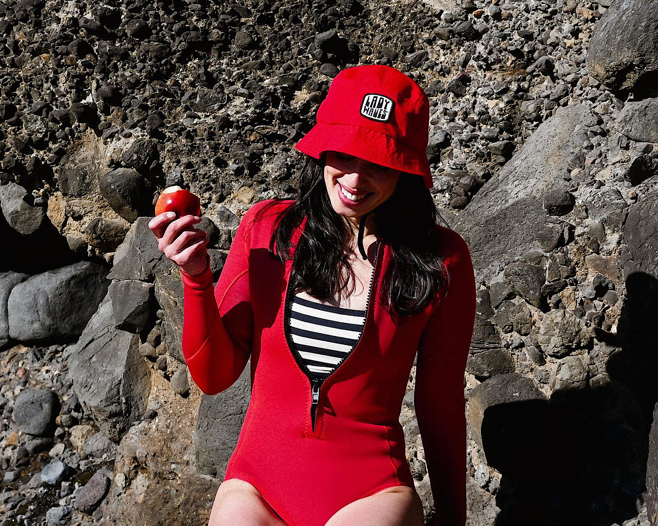 A woman wears a bright red wetsuit and red bucket hat as munches on an apple. 