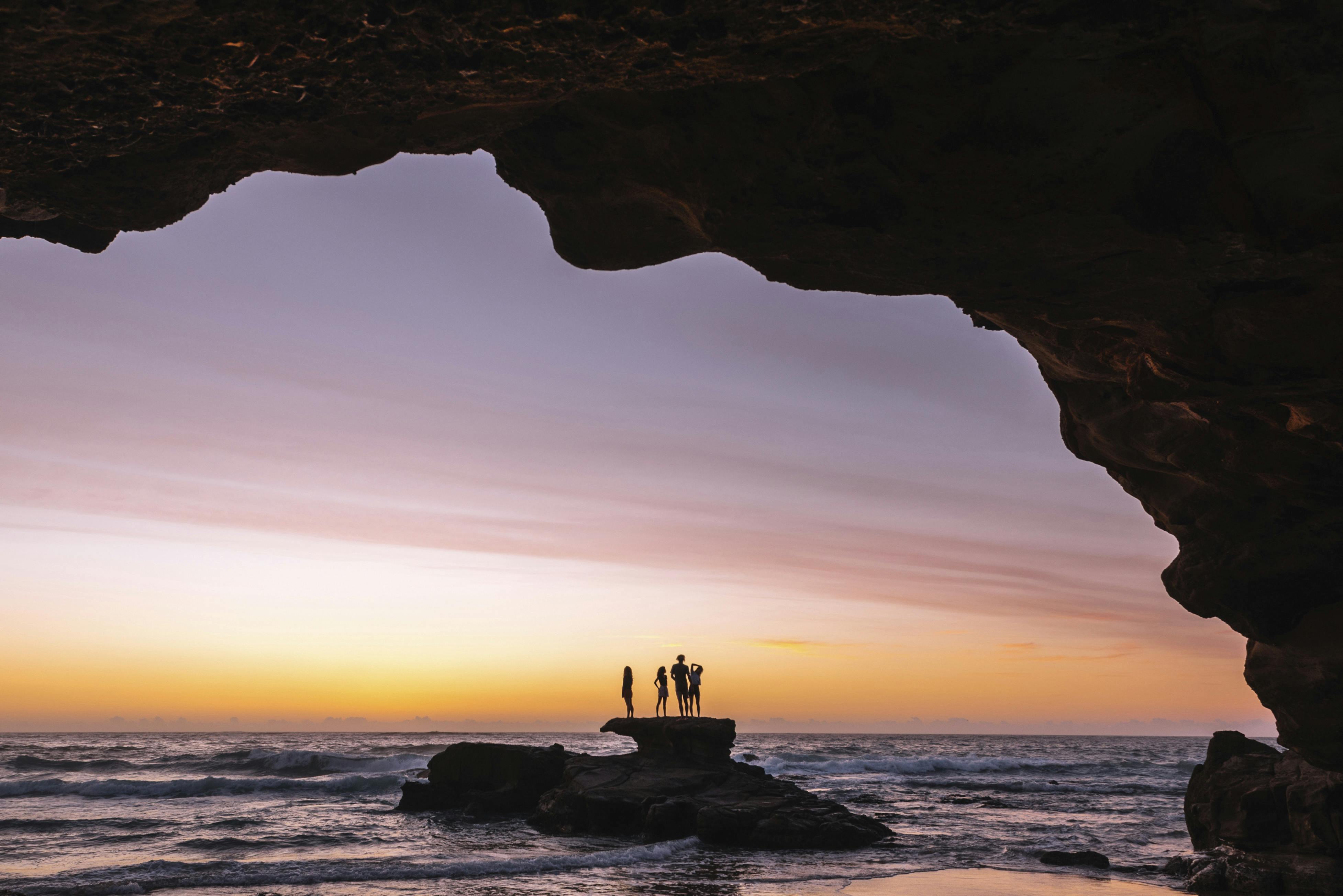 A group of friends stand on a rock just off the beach.