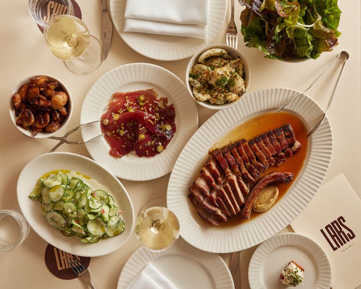 White table flat lay with plates filled with various foods: steak on the bone, roasted potatoes, cucumber salad, egg salad, charcuterie, lettuce, glasses of white wine.