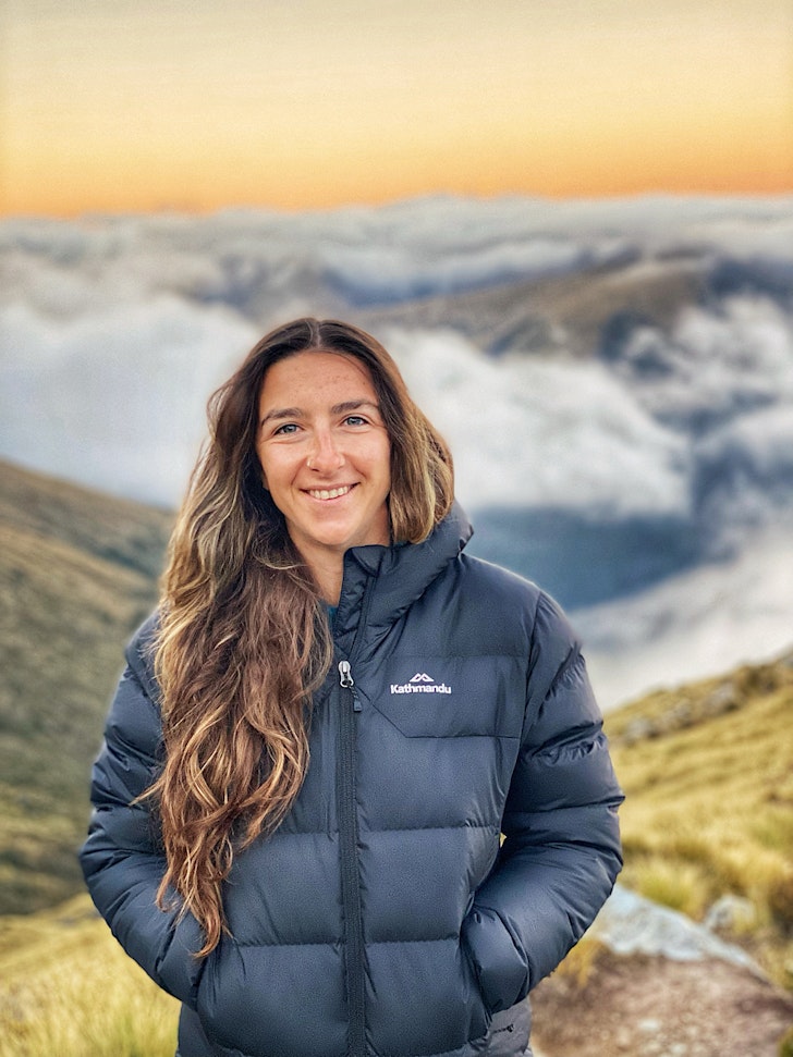 Lauren Sanderson wears a black puffer jacket and stands at the top of a stunning mountain. 