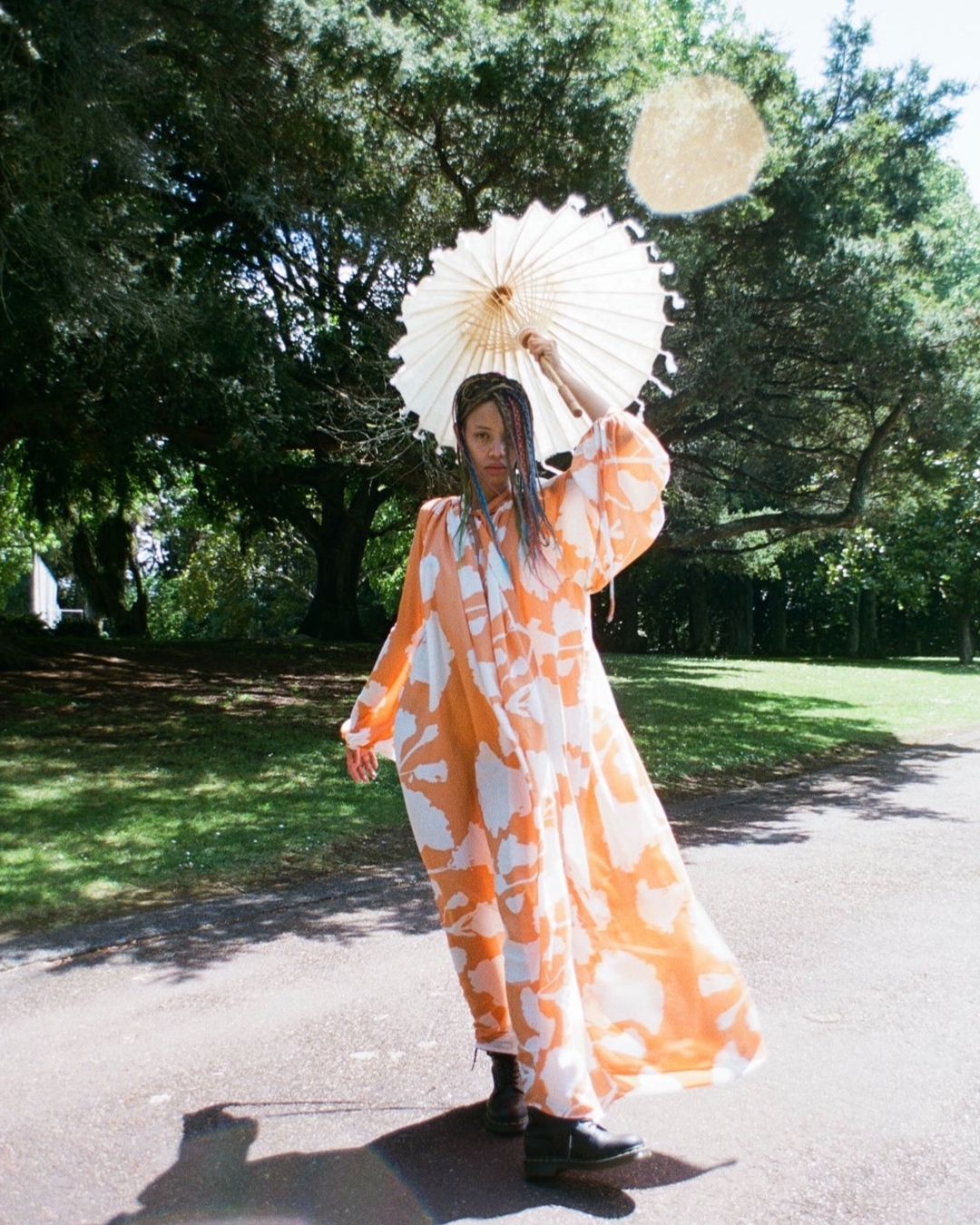 A woman with multicoloured braids, wearing an orange and white flowy dress holds a white parasol in a park. 