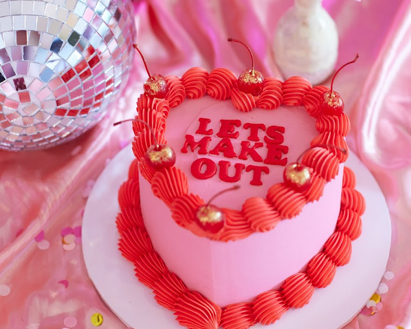 A vintage-inspired pink heart-shaped cake with the words ‘let’s make out’ iced on top of it, and a disco ball. 