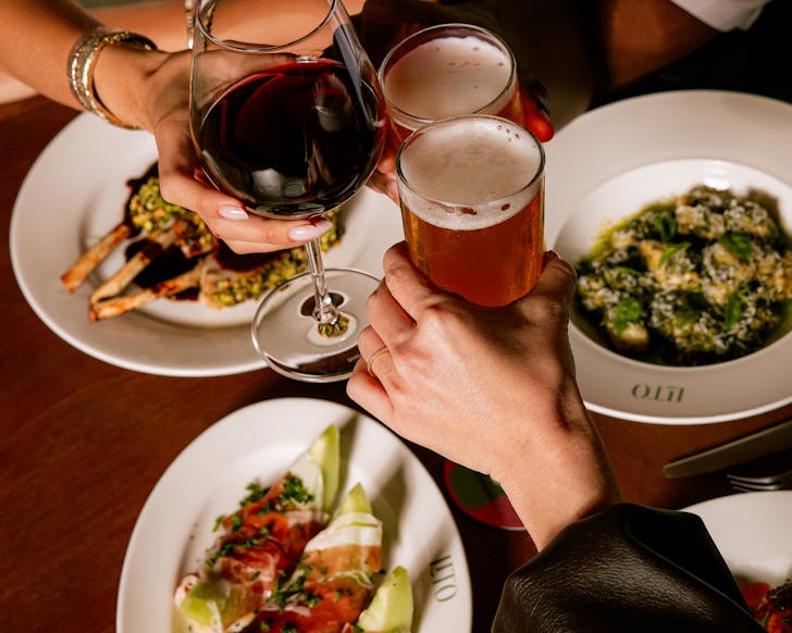 Hands clinking two beers and a glass of red wine over a table of food