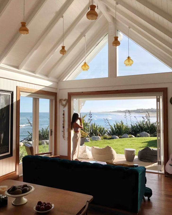 A woman looks out to sea from the living space at Little Church Bay Cottage one of the best beach house Airbnbs in NZ