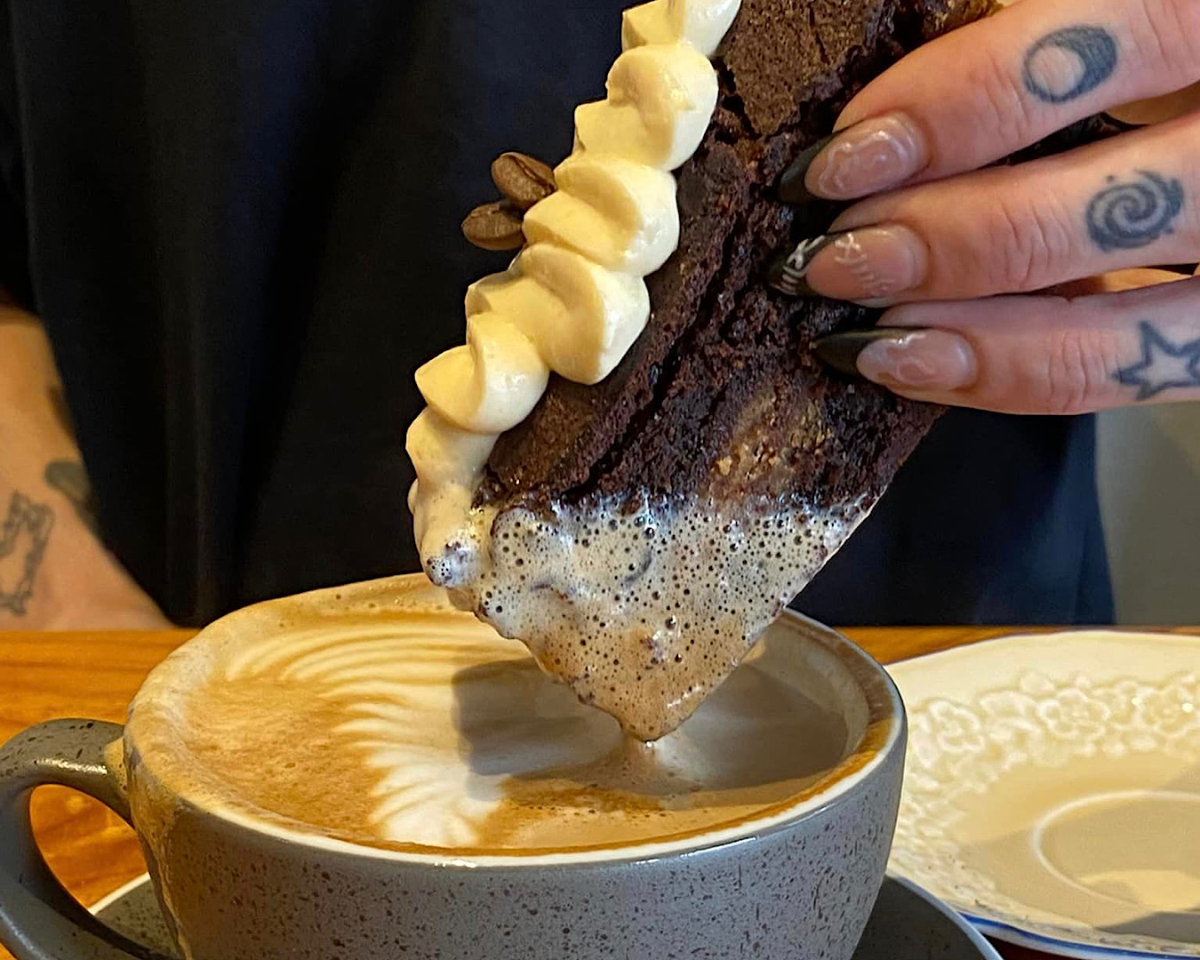 Someone with pretty nails dunks a slab of cake into their coffee, slightly overflowing the mug. 