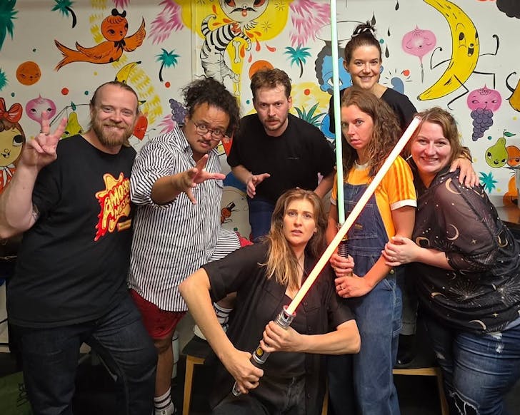 A group of comedians pose in front of a cartoon wall with light sabers. 
