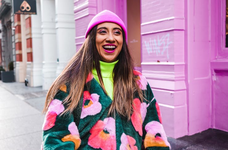 A woman wearing a colourful jacket stands in the street with a broad grin.