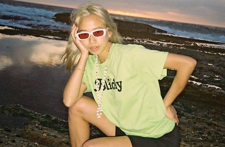 A woman crouches posing in front of the beach wearing white sleek sunglasses. 