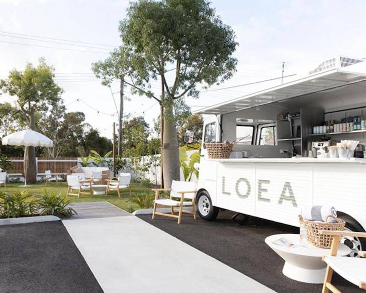 Shows sunny outdoor area with white chairs, lounges, leafy green plants and fairy lights. To the right you can see a white, outdoor food truck with 'LOEA' embossed on its front. Loea Boutique Hotel is one of the best accommodation spots in Maroochydore. 