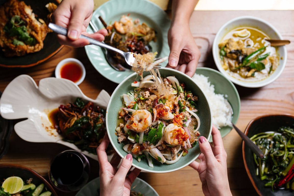 A table filled with food as two sets of hands pass a dish of prawns over the top