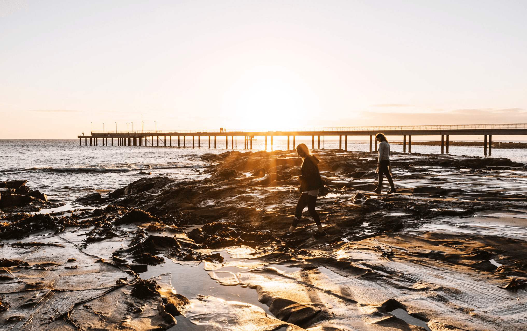 Two people at sunset walking across a rocky shore with a pier in the background at one of the best beaches in Victoria.