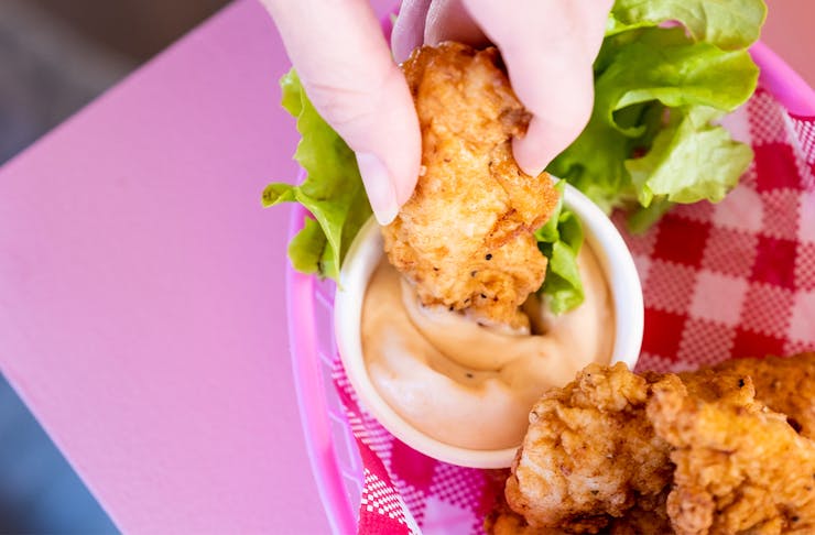 a fried chicken tender being dipped into a container of sauce on a pink table