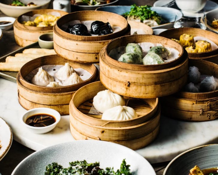 A table with many bamboo trays of dumplings in the centre. They are stacked on each other, and the dumplings are all different colours including the normal white along with green and black dumplings.