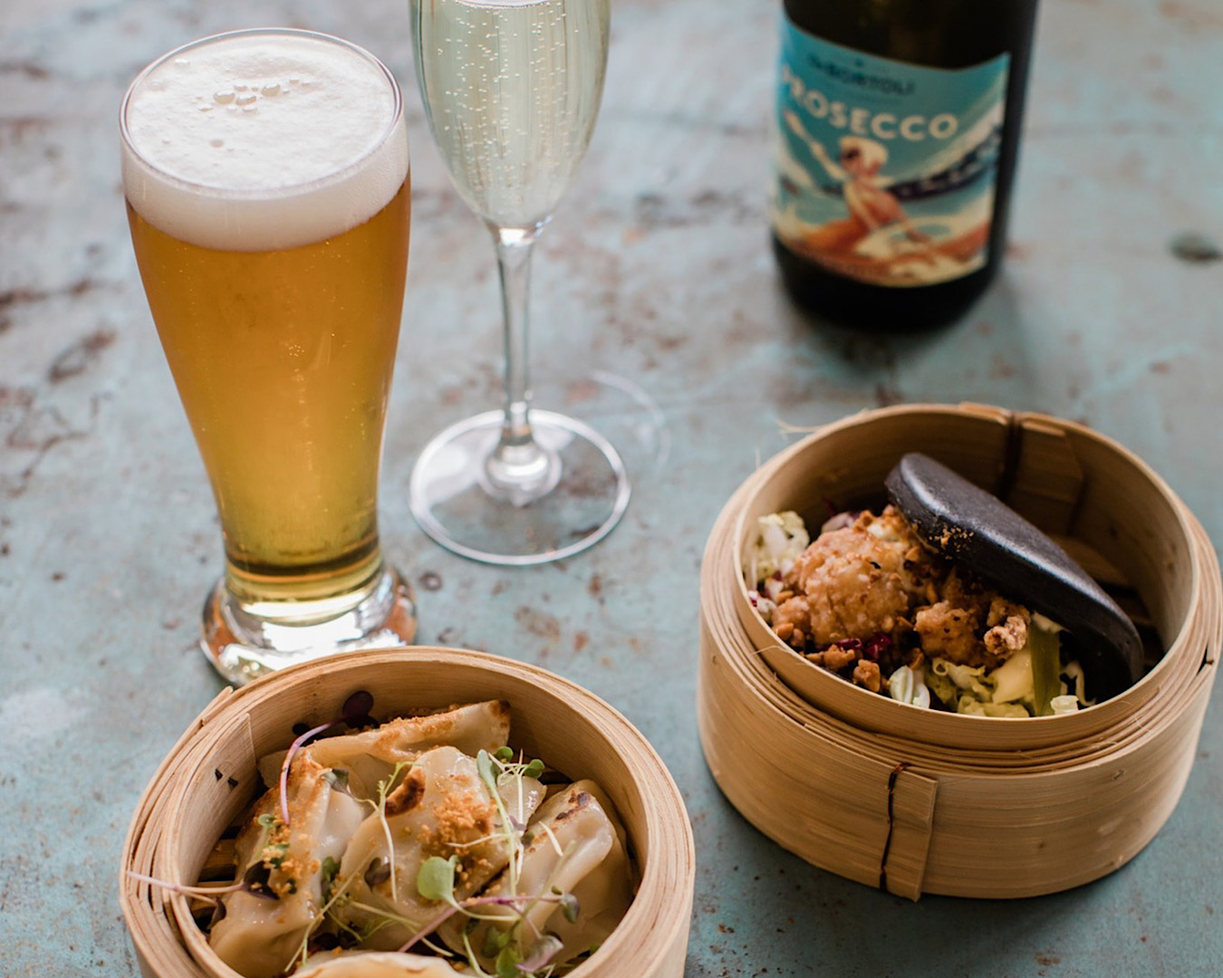 Two steamers with dumplings and bao sit on a table with a tall glass of beer and a flute of bubbles at Lula’s bottomless bao and bubbles. 