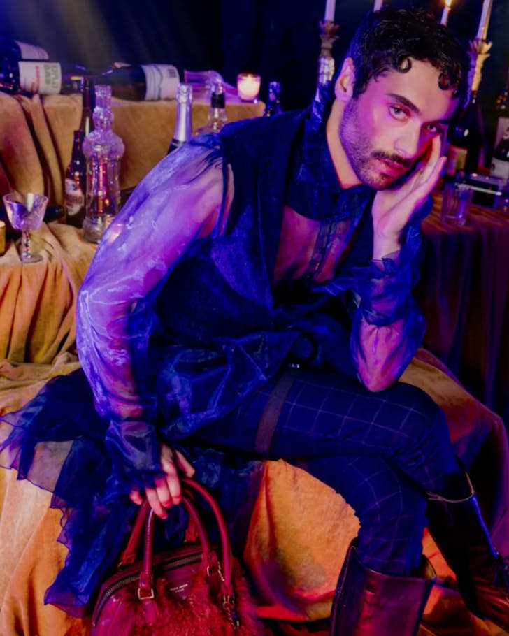 A cast member of MARY poses in racy and extravagant period costuming, holding a handbag. 