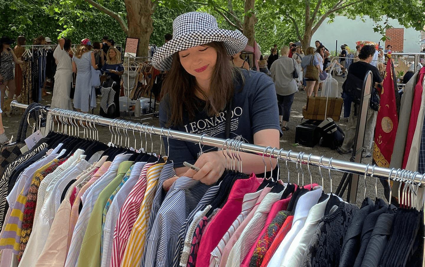 A woman in a black t shirt and checkered hat sorting through a rack of shirts at an outdoor market.