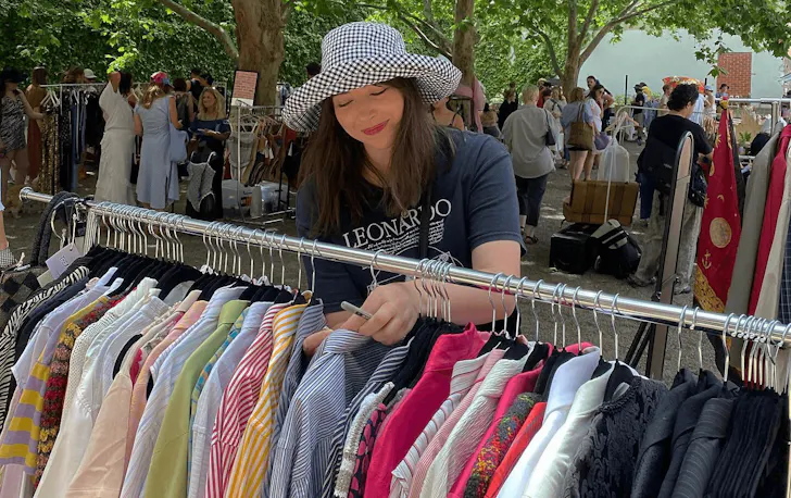 A woman in a black t shirt and checkered hat sorting through a rack of shirts at an outdoor market.