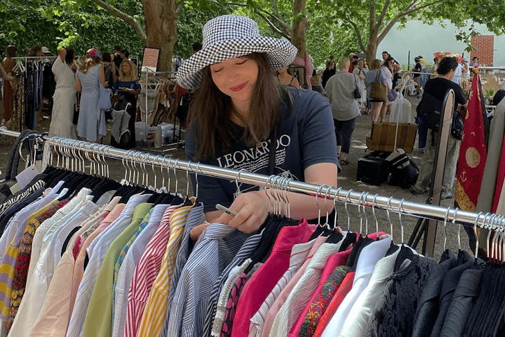 A woman in a black t shirt and checkered hat sorting through a rack of shirts at an outdoor market.